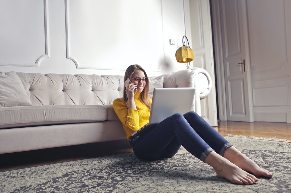 Color photo of a young woman sitting on the floor with back against sofa talking on a cell phone while looking at a laptop.