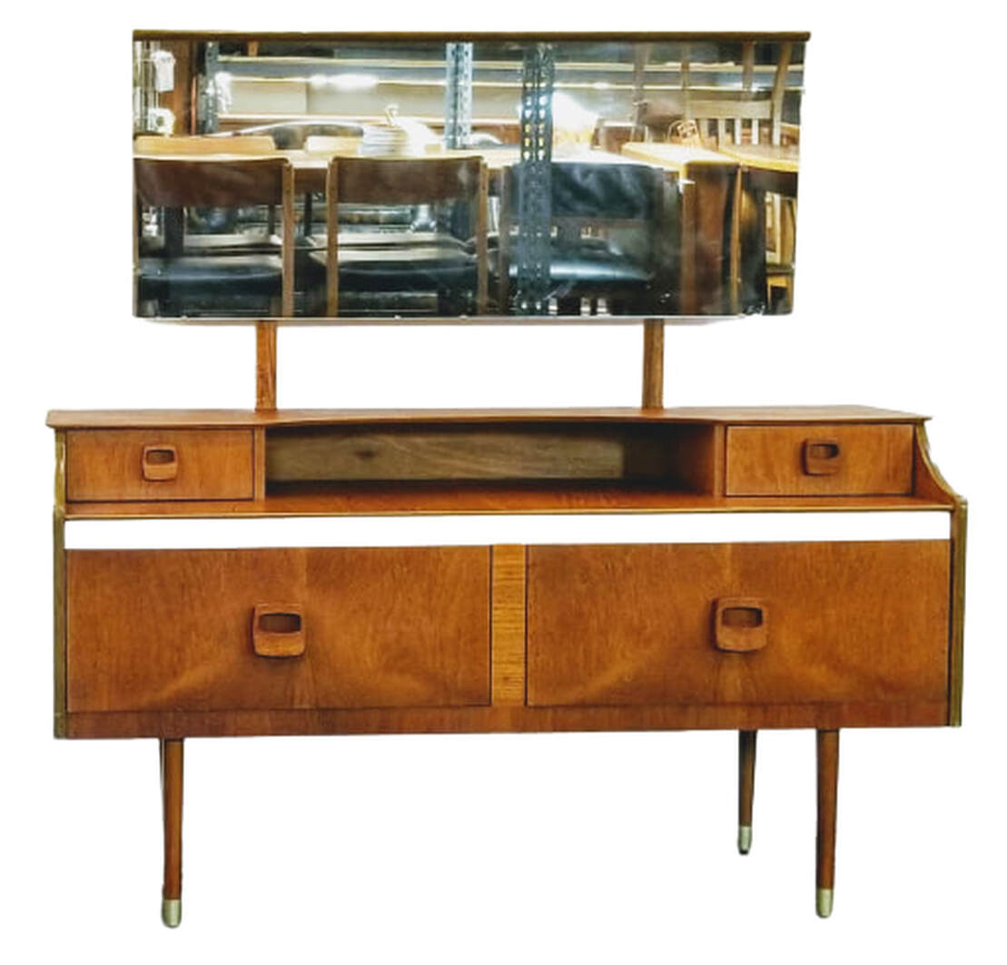 UK MCM bedroom vanity, or mirrored dressing table, features beautiful figured teak veneer and stylized wood pulls on the four storage drawers.  Across the center is a strip of white Melamine, a popular design addition for modern furniture in 1970s England.  The round tapered legs end in brass sabots.