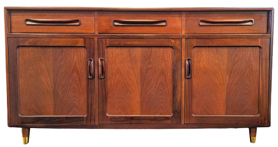 Fresco Buffet Board Base Unit 4075D was manufactured by E. Gomme Ltd. for their G-Plan brand in 1976.  Designed by Victor Bramwell Wilkins, this Fresco sideboard features three cupboards, each with one adjustable shelf, and three drawers lined and fitted for cutlery. The middle drawer has a safety latch.  The carcase is finished with crown-cut teak veneers and African afrormosia wood solids.  The drawers and cabinet doors feature the inset smiling pulls unique to G-Plan's Fresco range.  The carcase stands on four tapered feet with brass sabot.  56