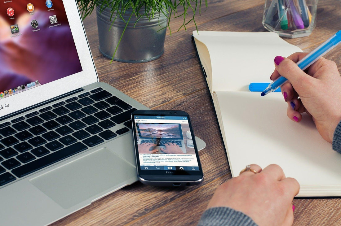 Woman wearing wedding ring preparing to write in journal with blue ink pen. Next to her hand on the desktop is a cel phone and a lap top computer.