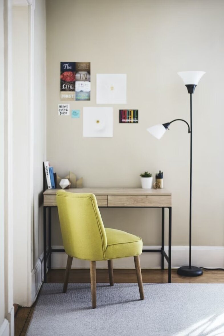 Color photo of a small corner home office with writing desk and bright green upholstered chair with splayed legs. A light grey carpet and a black pole lamp complete the scene.