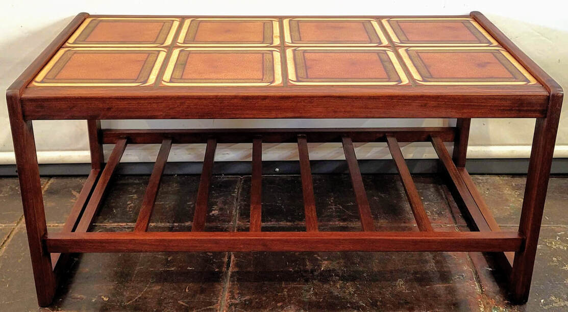 Rectangular teak wood coffee table with sled legs and a slatted magazine and book shelf.  The top is inset with ceramic tiles in orange, brown, and yellow colors.